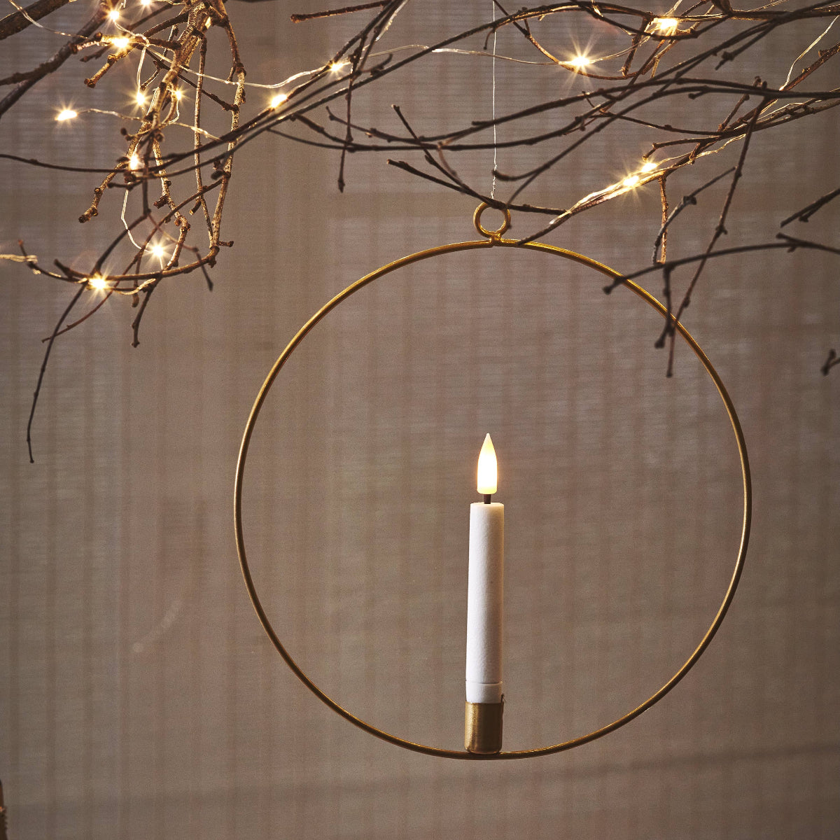 A gold circular metal wall hanging candle holder with a single white candle inside, suspended against a neutral textured wall with decorative string lights and star cutout ornaments.