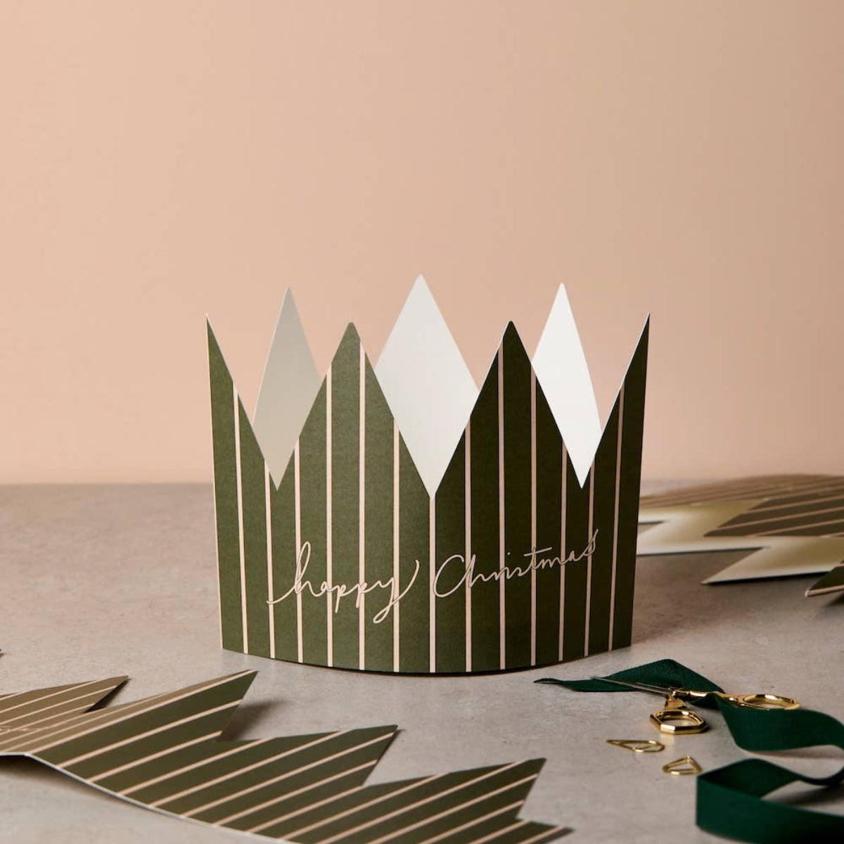 A green and white striped paper crown with 'Happy Christmas' written in gold script, displayed on a neutral surface with additional paper decorations in the background.