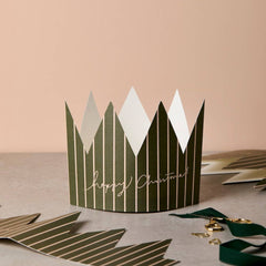 A green and white striped paper crown with 'Happy Christmas' written in gold script, displayed on a neutral surface with additional paper decorations in the background.