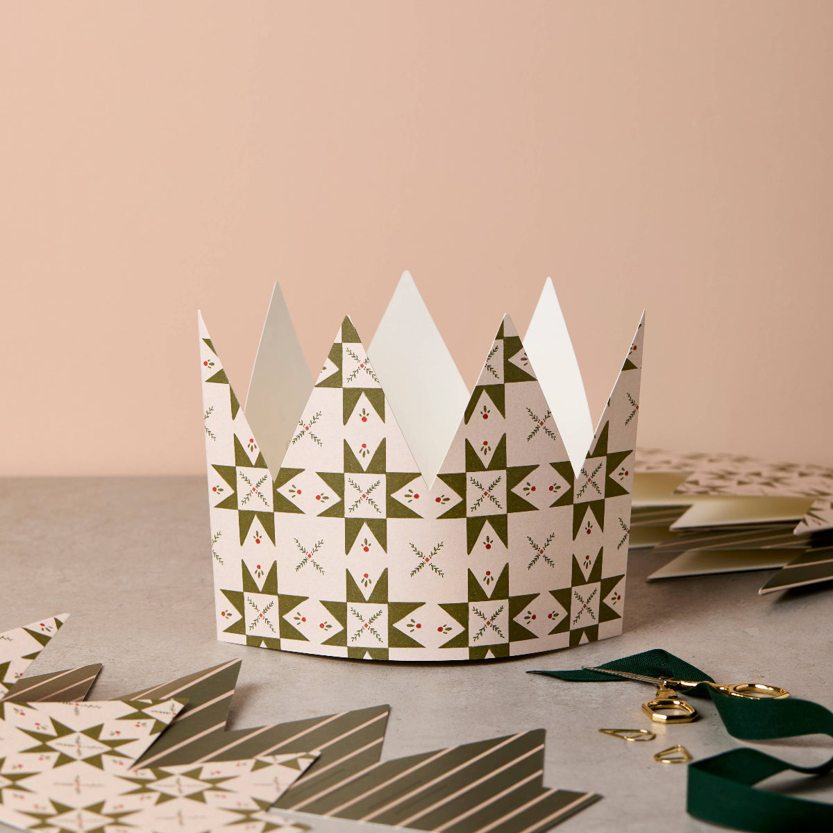 A paper crown party hat with a green and white geometric star pattern, displayed on a gray surface with other party hats and ribbons in the background.