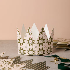 A paper crown party hat with a green and white geometric star pattern, displayed on a gray surface with other party hats and ribbons in the background.