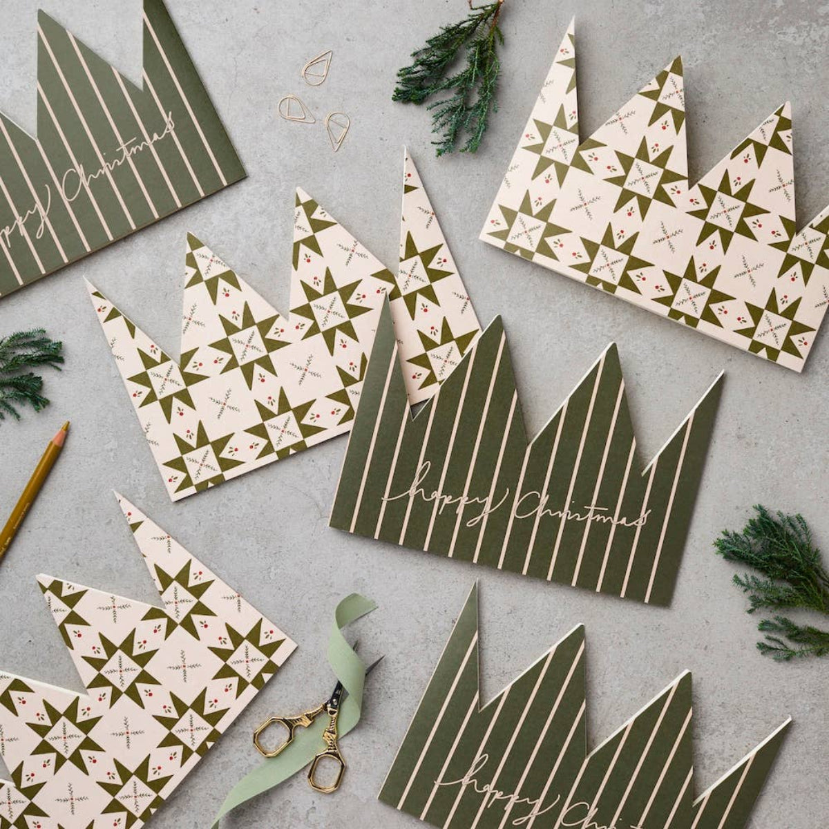 A flat-lay of Christmas crown-shaped paper decorations in olive green and cream, decorated with stripes, geometric star patterns, and 'Happy Christmas' script, arranged on a gray surface with scissors, ribbon, and evergreen sprigs.