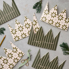A flat-lay of Christmas crown-shaped paper decorations in olive green and cream, decorated with stripes, geometric star patterns, and 'Happy Christmas' script, arranged on a gray surface with scissors, ribbon, and evergreen sprigs.