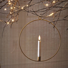 A gold circular metal wall hanging candle holder with a single white candle inside, suspended against a neutral textured wall with decorative string lights and star cutout ornaments.